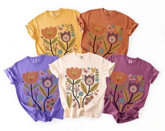 May include: Five short-sleeve t-shirts in yellow, orange, purple, cream, and dark pink. Each shirt features a graphic print of stylized flowers, leaves, and small birds in muted tones of orange, purple, green, and blue on the front center.