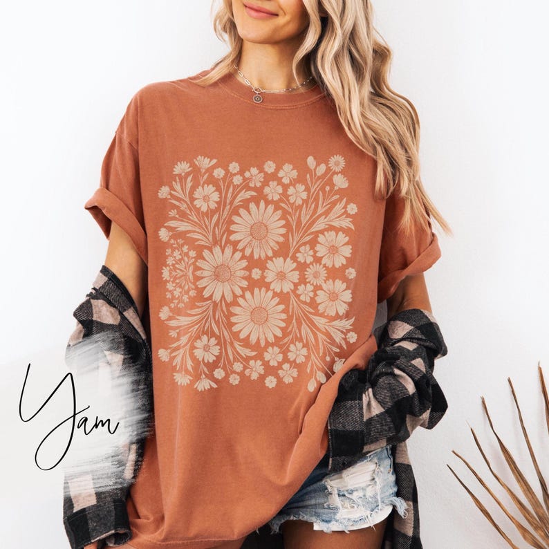 Pode incluir: Camiseta cor de ferrugem com um desenho floral com margaridas e folhas. O gr&aacute;fico est&aacute; centrado na frente da camiseta. As mangas est&atilde;o enroladas. A camiseta &eacute; combinada com shorts jeans e uma camisa xadrez preta e cinza.