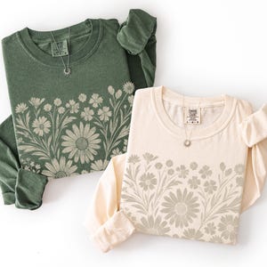 May include: Two long-sleeved shirts, one olive green and one cream, each featuring a floral design across the chest. Both shirts have a crew neck and a small silver necklace. The floral design includes daisies and other flowers.