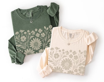 May include: Two long-sleeved shirts, one olive green and one cream, each featuring a floral design across the chest. Both shirts have a crew neck and a small silver necklace. The floral design includes daisies and other flowers.