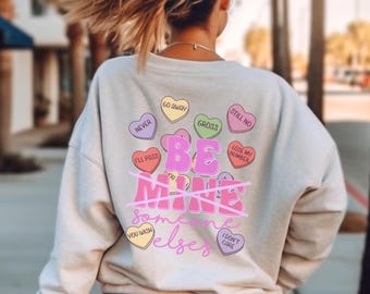 May include: A light beige sweatshirt with a pink graphic that says "Be Mine" with a line through it and the words "someone else" below. The graphic is surrounded by colorful candy hearts with phrases like "Go Away", "Still No", "Gross", "Never", "I'll Pass", "Lose My Number", "You Wish", and "I Don't Care".