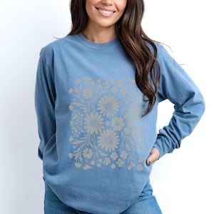 May include: A long-sleeved, blue shirt featuring a floral design. The floral pattern is in a lighter shade of beige and includes daisies and other flowers. The shirt is made of a soft material and has a relaxed fit.