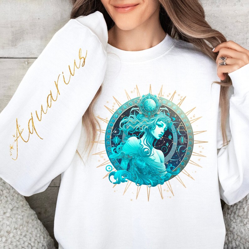 Puede incluir: Sudadera blanca con una ilustraci&oacute;n dorada y azul de una mujer con el pelo suelto y una corona. La ilustraci&oacute;n est&aacute; rodeada de un c&iacute;rculo de estrellas doradas y la palabra "Aquarius" est&aacute; escrita en letras doradas en la manga.
