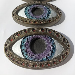 Puede incluir: Tres objetos decorativos con forma de ojo con un marco dorado y detalles intrincados. Los ojos tienen un iris morado con un anillo exterior azul y blanco.