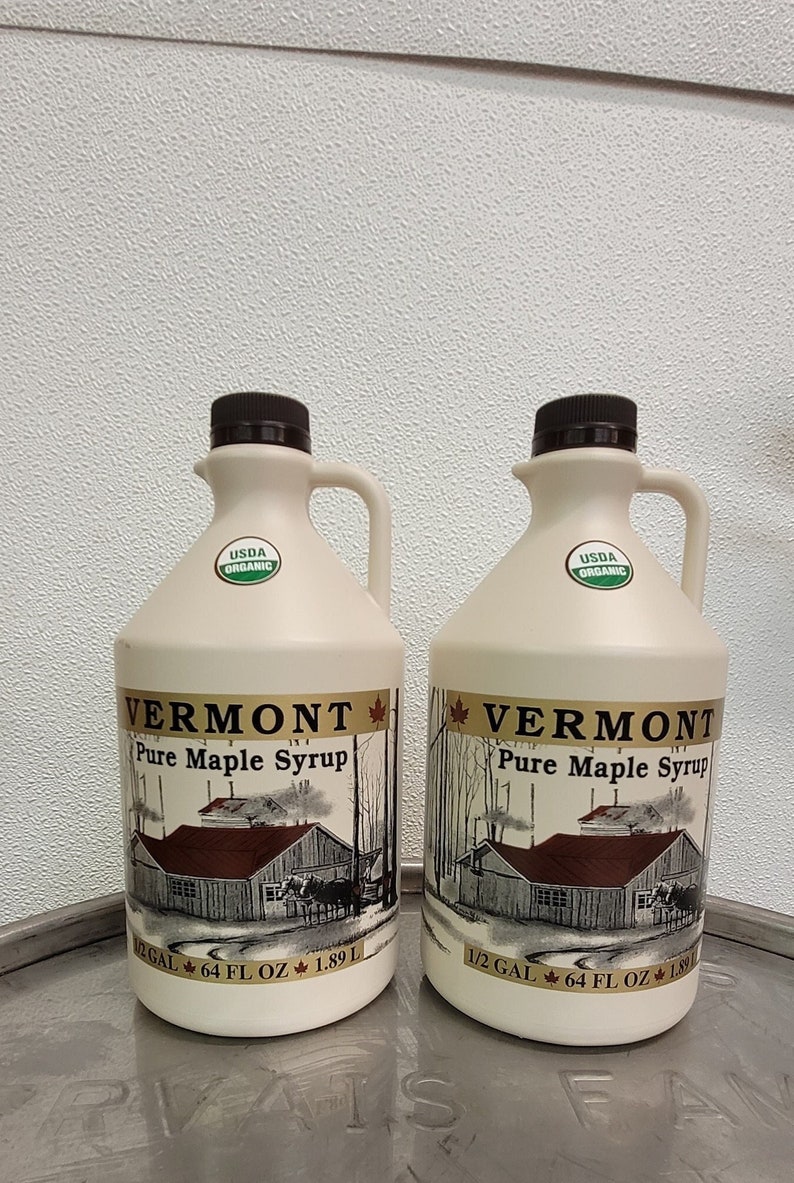 Pu&ograve; includere: Due taniche da 1,89 litri di sciroppo d'acero puro del Vermont, certificate USDA Organic. Le taniche sono bianche con un tappo marrone e un'etichetta che raffigura una casa con tetto rosso e una foglia d'acero.