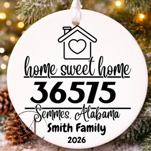 May include: A round white ceramic ornament featuring a black line drawing of a house with a heart, the text home sweet home, the zip code 36575, Semmes, Alabama, Smith Family, and the year 2026, suspended by a white ribbon for holiday display.