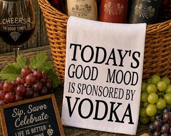 May include: A rustic scene with a wine theme. A wicker basket holds three wine bottles, a red and black plaid cloth, and a white towel with the text "TODAY'S GOOD MOOD IS SPONSORED BY VODKA." A wine glass, grapes, and a sign that reads "Wine Festival" are also present.