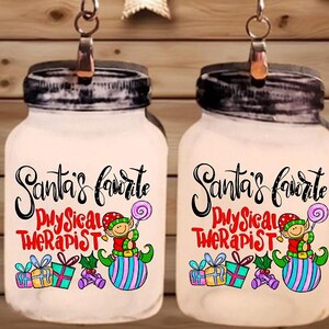 May include: A pair of Christmas-themed earrings. Each earring features a small, white jar with a black lid, adorned with the text "Santa's Favorite Physical Therapist" and a cartoon elf holding a lollipop. The background includes wooden planks and star-shaped lights.