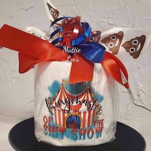 Puede incluir: Un regalo cilíndrico envuelto en celofán transparente, adornado con cintas rojas, blancas y azules. El regalo presenta un diseño de carpa de circo con el texto "Ringmaster of the Shit Show". La cinta incluye emojis de caca sonrientes.