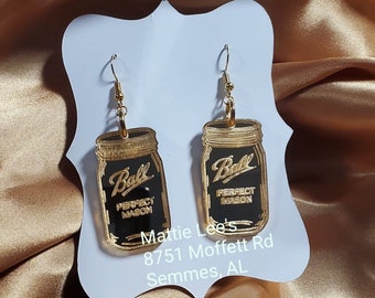 May include: Gold-colored earrings shaped like Ball brand mason jars with black glass and gold lettering that reads "Ball Perfect Mason". The earrings are hanging from a white card with the text "Mattie Lee's 8751 Moffett Rd Semmes, AL".