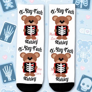 May include: White socks with a black toe and heel, featuring a brown teddy bear holding an X-ray. The text "X-Ray Tech" and the name "Ashley" are printed on the socks. The background includes medical-themed illustrations.