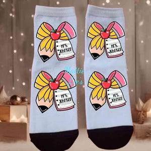 May include: A pair of light grey socks with a black toe and heel. Each sock features a pencil design with a red apple, a pink ribbon, and a notepad that reads "Mrs. Johnson".