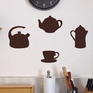 Peut inclure: Un ensemble de quatre autocollants de cuisine en silhouette brune, comprenant une théière, une bouilloire, une cafetière et une tasse à thé et sa soucoupe. Les autocollants sont sur un mur blanc.