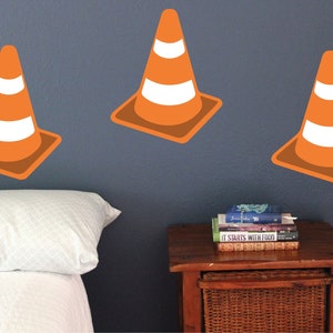 May include: Three orange and white traffic cone wall decals on a blue wall. The decals are in a row, with the middle one slightly higher than the others. There is a white bed with a white pillow in the foreground.