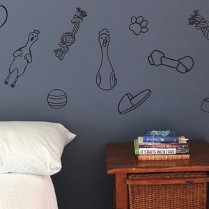 Op de afbeelding: Een slaapkamer met een wit bed en een houten nachtkastje. De muur is versierd met zwart-witte wanddecoraties met een hondenmotief, waaronder een bot, een bal, een hondenspeelgoed, een hondenkom en een pootprint. Het nachtkastje heeft een stapel boeken erop, waaronder een met de titel "It Starts With Food".