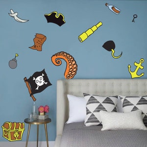 May include: A blue wall with a variety of pirate-themed wall decals, including a skull and crossbones flag, a treasure chest, an octopus tentacle, a spyglass, a bottle with a message, a pirate hat, a key, a hook, and an anchor. The decals are on a wall behind a bed with a white headboard and gray bedding.