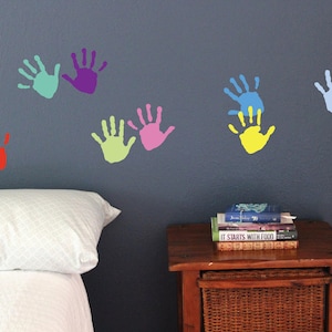 May include: A blue wall with colorful handprint decals in various shades of blue, green, purple, pink, and orange. The handprints are arranged in a row across the wall. There is a white bed with a white pillow in the foreground.