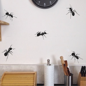 May include: Five black ant silhouettes are displayed on a white wall. The ants are in various positions, some facing left and some facing right. The ants are all the same size and shape.
