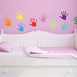 May include: A white daybed with a pink bedspread and a pink pillow. The wall behind the bed is pink and has colorful handprint decals in various colors, including blue, yellow, red, pink, green, and purple.