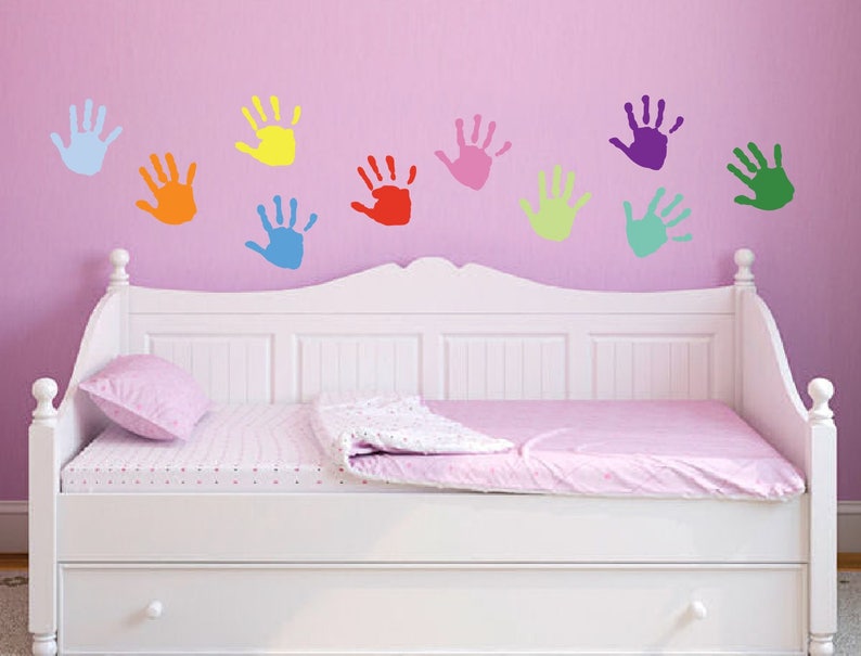 May include: A white daybed with a pink bedspread and a pink pillow. The wall behind the bed is pink and has colorful handprint decals in various colors, including blue, yellow, red, pink, green, and purple.