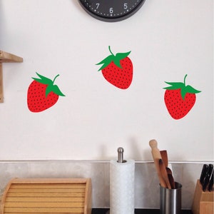 Puede incluir: Tres pegatinas de pared de fresa roja con hojas verdes en una pared blanca.