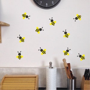 May include: A white wall with a black clock and multiple yellow and black bee decals. The wall is in a kitchen with a white countertop and a wooden bread box.
