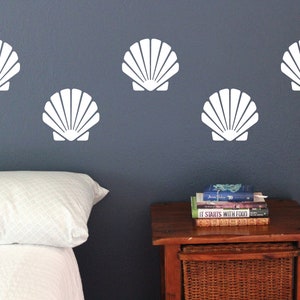 May include: Five white seashell wall decals on a dark blue wall above a white bed with a white patterned pillow. A wooden nightstand with a stack of books is to the right of the bed.