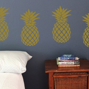 May include: Four gold pineapple wall decals on a blue wall above a white bed with a white pillow and a wooden nightstand with a stack of books on top.