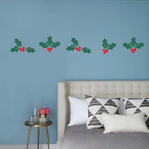 May include: Green holly leaf and red berry wall decals arranged in a horizontal line against a light blue wall. The decals are above a bed with white and patterned pillows. A small gold side table with a glass bottle and flowers is visible.