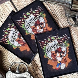 May include: Two rectangular fabric patches featuring a graphic of a cartoon character holding a white globe with the text "ROCK 'N' ROLL" above it, positioned between two silver-colored toy revolvers with ornate engraved patterns on the barrels.