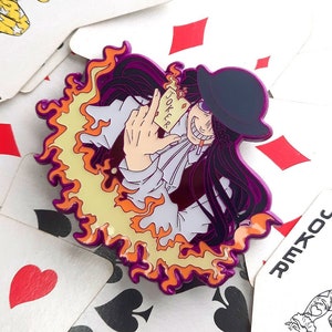 Puede incluir: Un pin de esmalte morado y amarillo con un personaje con un sombrero de copa y un diseño de llama. El personaje sostiene una tarjeta que dice "Joker". El pin está rodeado de cartas de juego.