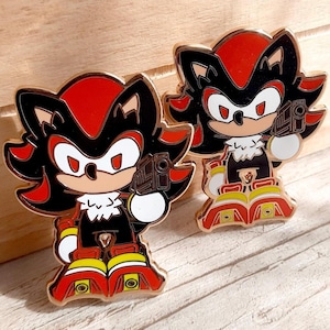 May include: Two gold-coloured enamel pins featuring a cartoon character with red and black fur, holding a gun. The character is wearing a red and yellow outfit.
