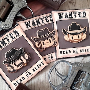 Puede incluir: Tres carteles de "Se busca" con ilustraciones de vaqueros de dibujos animados y el texto "Dead or Alive" se muestran cerca de dos revólveres plateados. Los carteles tienen una estética vintage con una combinación de colores marrón y negro, y los pines de vaquero tienen un sombrero negro.