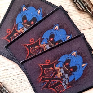 Könnte beinhalten: Drei quadratische Aufnäher mit einer blauen Cartoonfigur mit roten Augen und einem finsteren Grinsen. Die Aufnäher sind auf einer Holzoberfläche gestapelt, mit zwei silbernen Revolvern im Hintergrund.