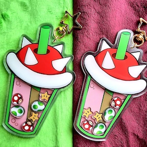 May include: Two clear plastic charms shaped like a boba tea cup with a green straw and a red and white mushroom top. The charms are filled with green and yellow star-shaped candies and red and green mushroom-shaped candies. The charms are attached to gold keychains with star-shaped charms.