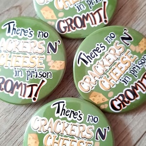 Könnte beinhalten: Vier grüne runde Buttons mit dem Text "There's no CRACKERS N' CHEESE in prison GROMIT!" in Schwarz, Weiß und Beige. Die Buttons haben eine Käse- und Cracker-Grafik. Die Buttons liegen auf einer Holzoberfläche.