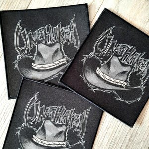 May include: Three black square patches with a white graphic of a cowboy hat and the word "UNSHAKEN" in a stylized font. A decorative silver revolver is in the background.