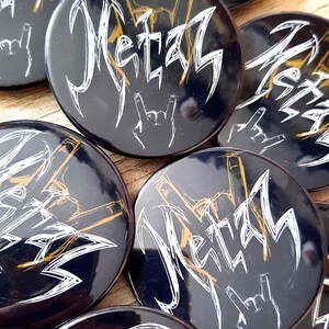 Puede incluir: Un conjunto de botones redondos negros con la palabra "Metal" en una fuente blanca estilizada. Cada botón presenta un gesto con la mano y detalles dorados. Los botones están dispuestos sobre una superficie de madera.