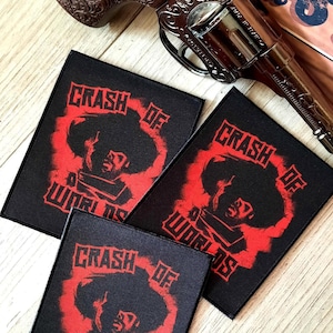 Op de afbeelding: Drie zwarte patches met de tekst "CRASH OF WORLDS" in het rood, met een gestileerde afbeelding van een persoon. Een decoratief pistool is boven de patches geplaatst.