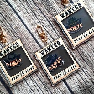 May include: Three gold keychains with a cartoon character wearing a cowboy hat and a wanted poster design. The poster says "WANTED" and "DEAD OR ALIVE".