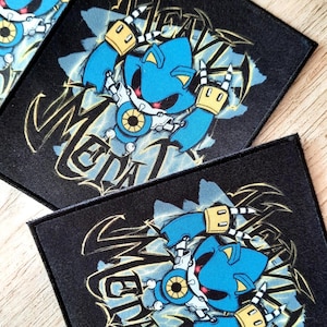 May include: Black square patch featuring a blue robot character with the word "METAL" in a stylized font. The robot has yellow accents and is set against a blue lightning background. The patch is likely for clothing or accessories.