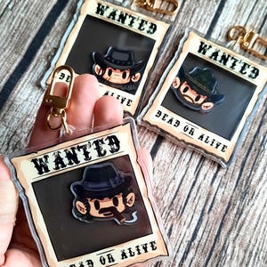 May include: Three clear plastic keychain charms with a cartoon cowboy character in a black hat and a mustache. Each charm has a gold metal ring and the text "WANTED DEAD OR ALIVE".