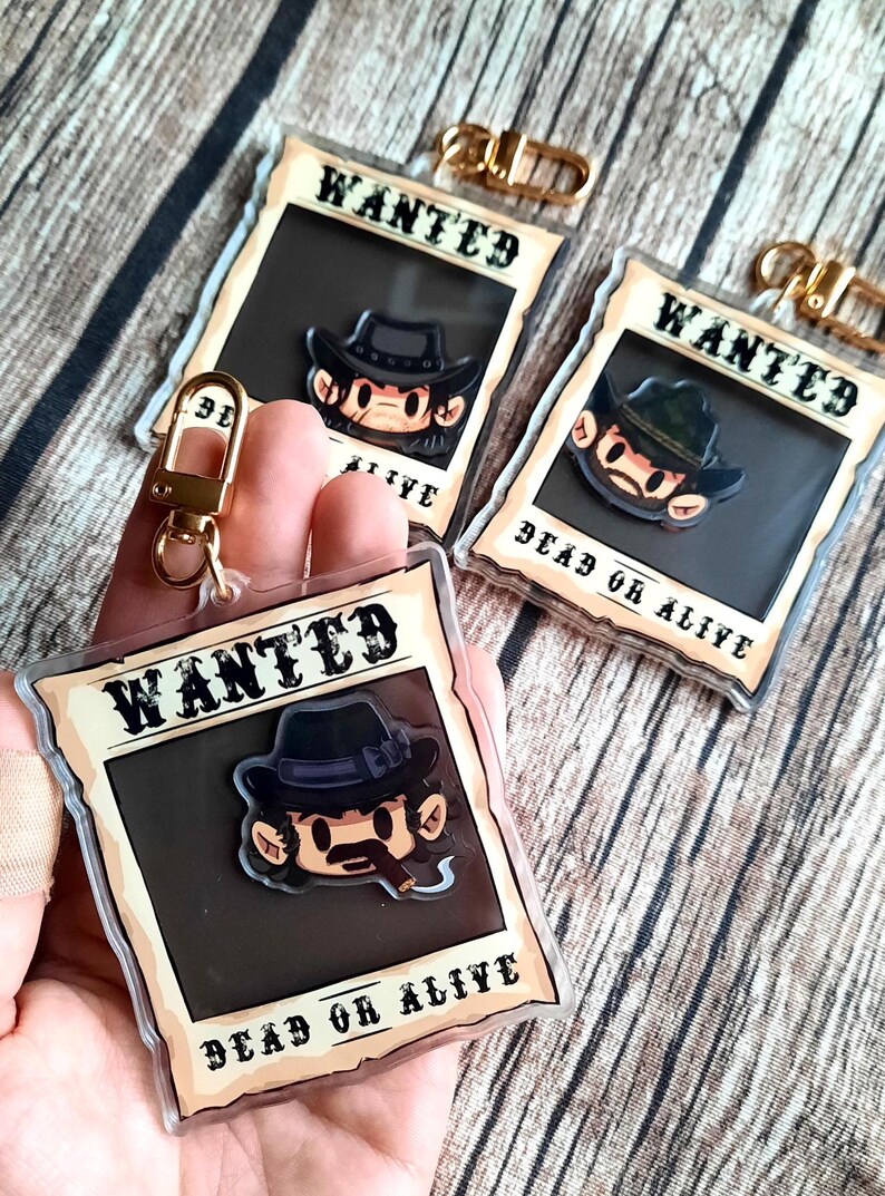May include: Three clear plastic keychain charms with a cartoon cowboy character in a black hat and a mustache. Each charm has a gold metal ring and the text "WANTED DEAD OR ALIVE".