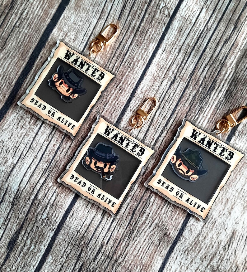 May include: Three gold keychains with a cartoon character wearing a hat and a mustache. Each keychain has a white background with the word "WANTED" in black text and the words "DEAD OR ALIVE" in black text.