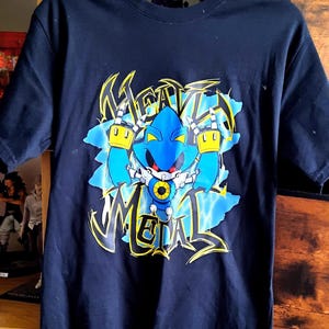 May include: Navy blue t-shirt featuring a blue robot character with yellow accents and the text "HEAVY METAL" in a graffiti-style font. The graphic is set against a light blue background with lightning bolt designs.