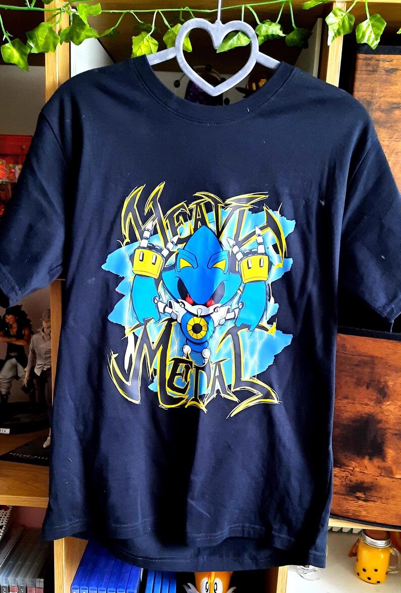 May include: Navy blue t-shirt featuring a blue robot character with yellow accents and the text "HEAVY METAL" in a graffiti-style font. The graphic is set against a light blue background with lightning bolt designs.