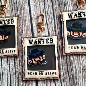 May include: Three clear acrylic keychains with a cartoon character wearing a cowboy hat and a wanted poster design. The text on the poster says "WANTED DEAD OR ALIVE".