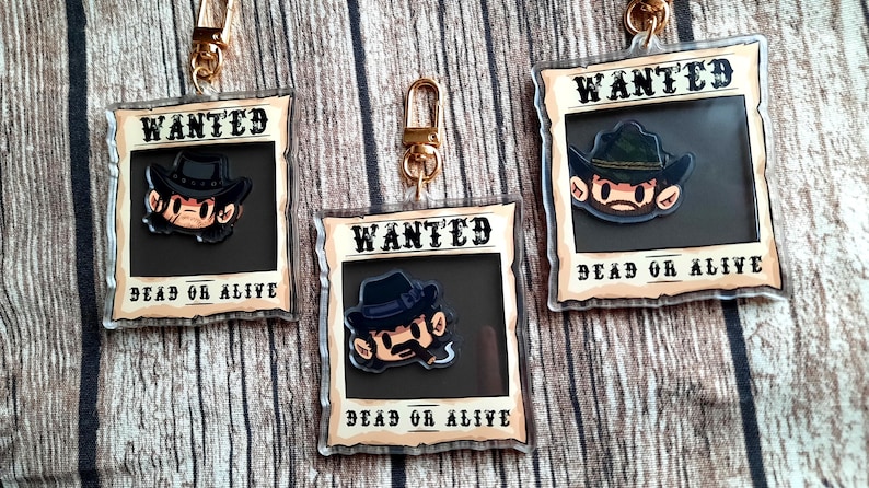 May include: Three clear acrylic keychains with a cartoon character wearing a cowboy hat and a wanted poster design. The text on the poster says "WANTED DEAD OR ALIVE".