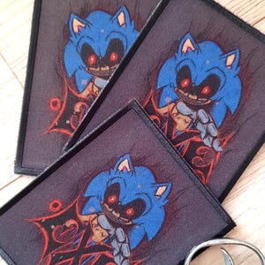 May include: Three rectangular patches featuring a blue cartoon character with red eyes and a sinister expression. The patches are on a dark background with red accents. Two silver toy revolvers are in the background.