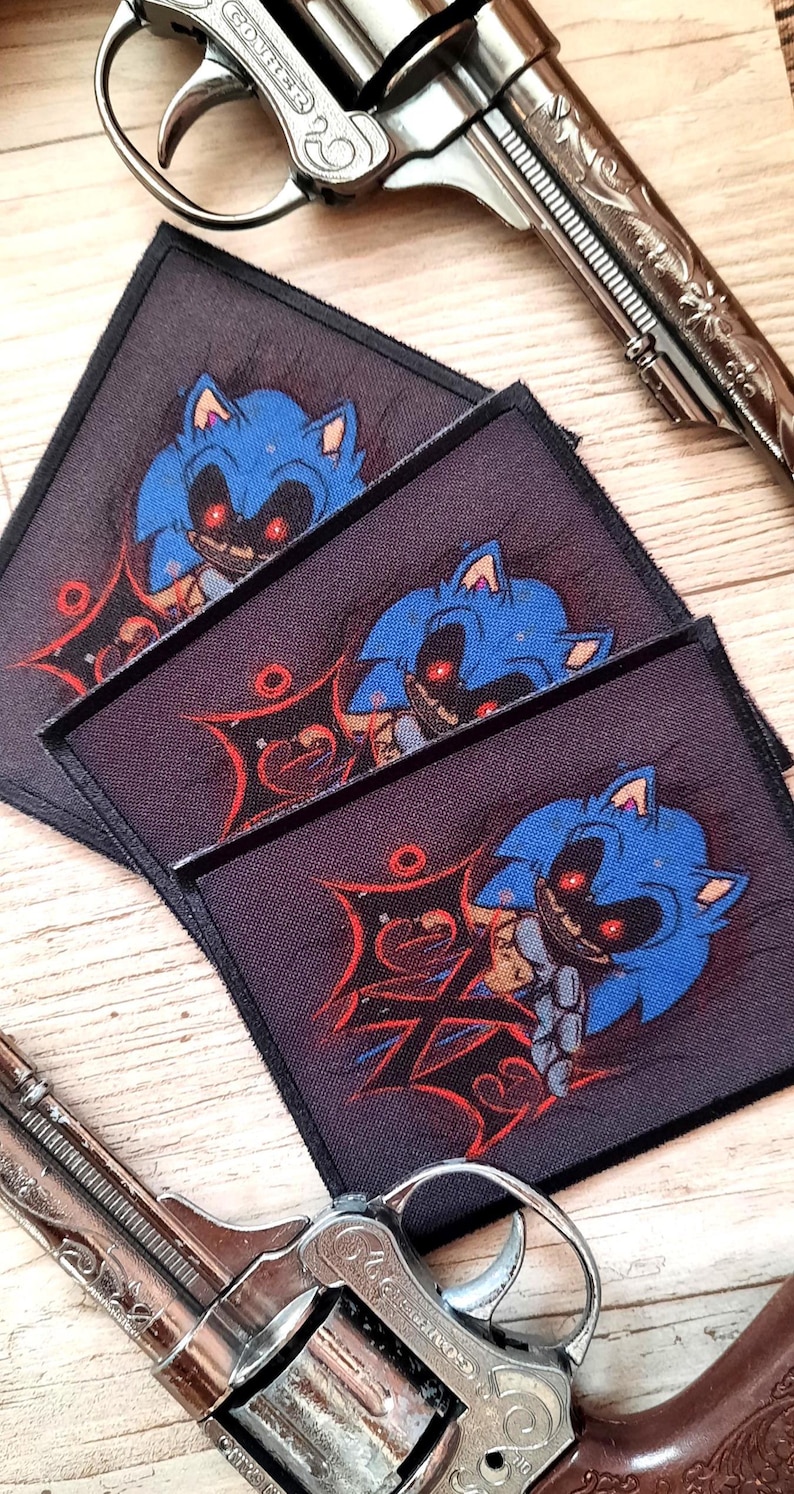 May include: Three square patches featuring a blue cartoon character with red eyes and a sinister grin. The patches are stacked on a wooden surface, with two silver revolvers in the background.